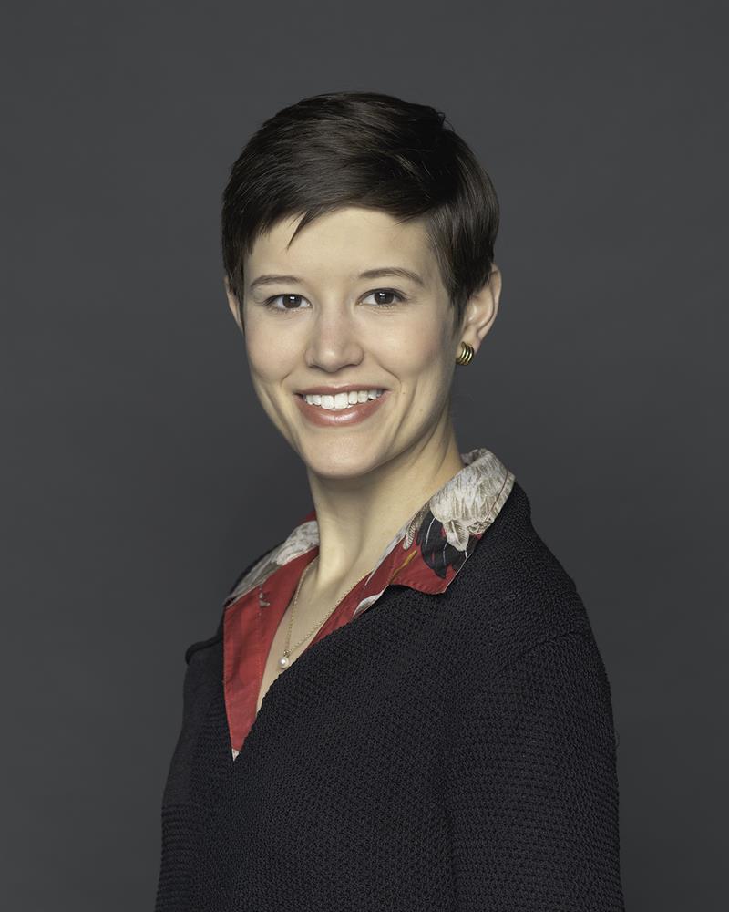 A woman with short hair, wearing a multicolored shirt and dark jacket.