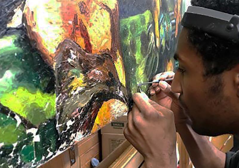 Close-up image of a student, wearing a magnifying visor and working with a small brush, applying consolidant to flaking paint on a painting.