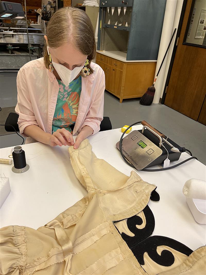 A student sits at a table and hand-stitches lining fabric to the dress sleeve.