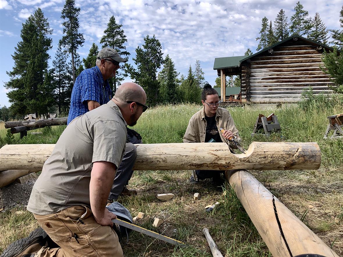 A student uses a chisel to carve a notch in a log while two teachers watch.