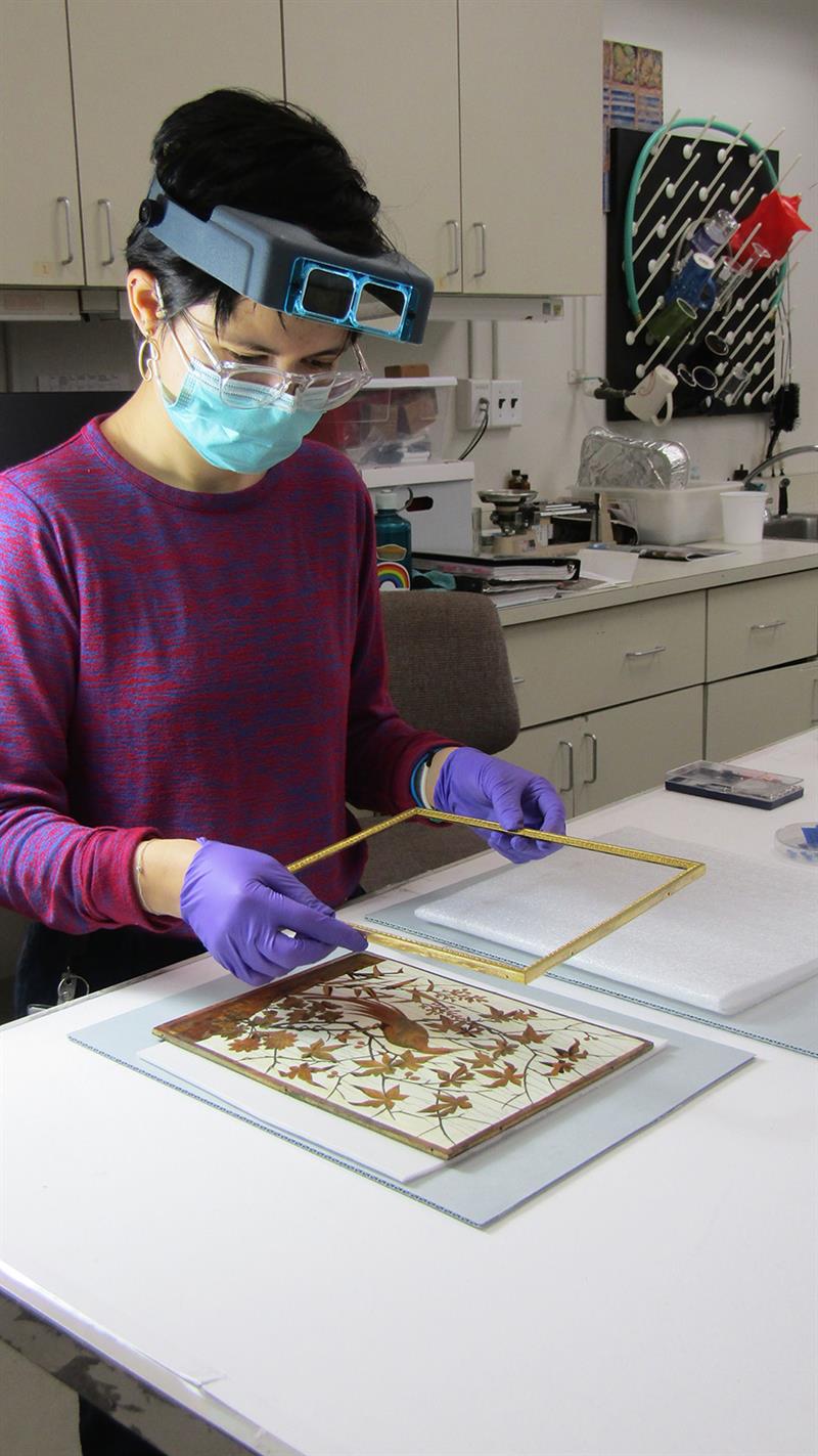 A student, wearing a magnifying visor and gloves, places a frame on a marquetry panel.